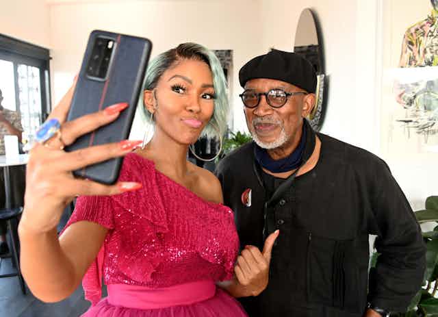 A stylish woman with dyed hair and a bright pink dress takes a selfie with an older man dressed in black with a grey beard, round spectacles and beret.