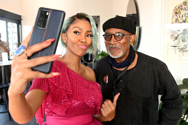 A stylish woman with dyed hair and a bright pink dress takes a selfie with an older man dressed in black with a grey beard, round spectacles and beret.