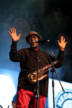 A musician performs into a microphone, his hands up and fingers splayed, a saxophone around his neck.