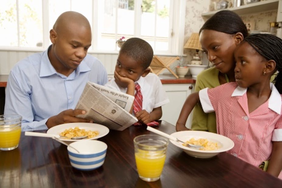 A young husband and wife share the newspaper with their son and daughter in school uniform while having cereal and orange juice for breakfast. The boy is covering his mouth with his hand in shock