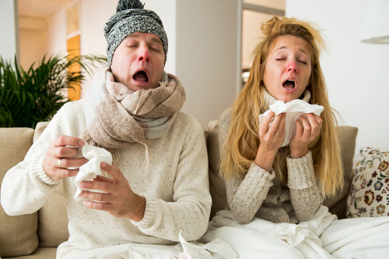 Un couple éternuant et se mouchant, effondré sur son canapé.