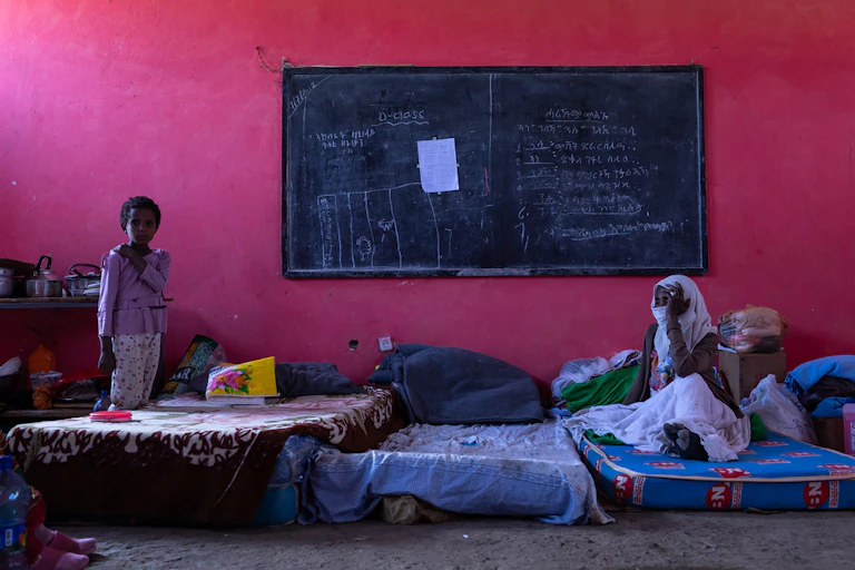 A woman sits on a matress under a blackboard with a girl standing near her.