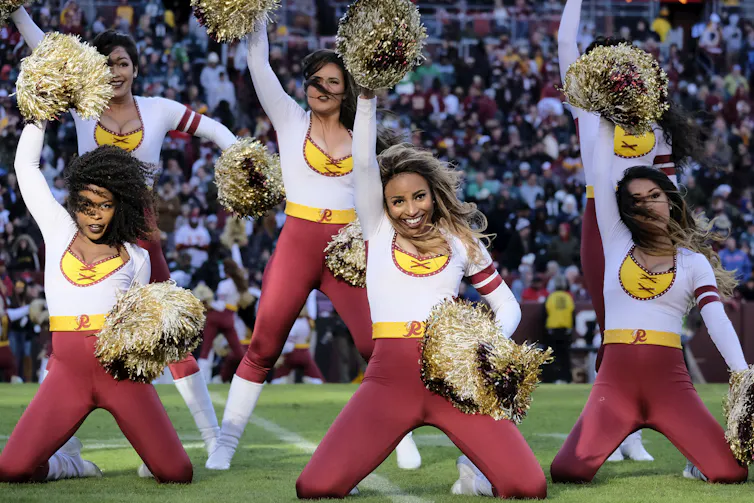 Six Washington Football Team cheerleaders perform on the field. The stands are full behind them. The girls are wearing red spandex pants, white tops and red and yellow pompoms.