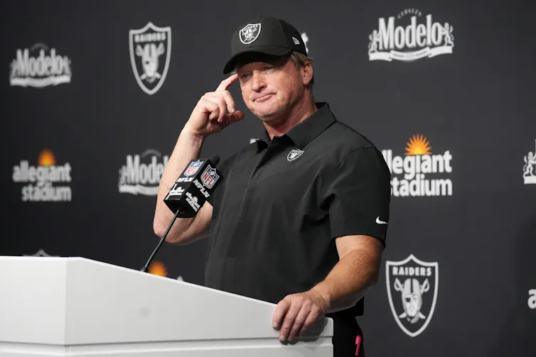 Former Las Vegas Raiders coach Jon Gruden stands behind a podium. He is wearing all black and has a perplexed look on his face. He is scratching his temple with one of his fingers.