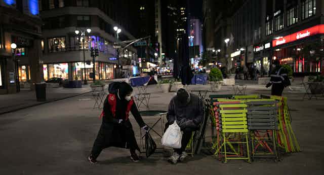 A woman wearing a mask hands a bag of food to a homeless man.