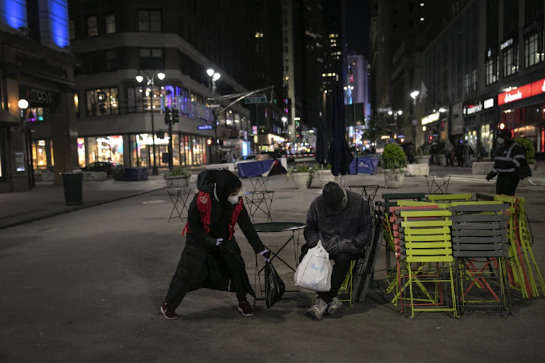 A woman wearing a mask hands a bag of food to a homeless man.
