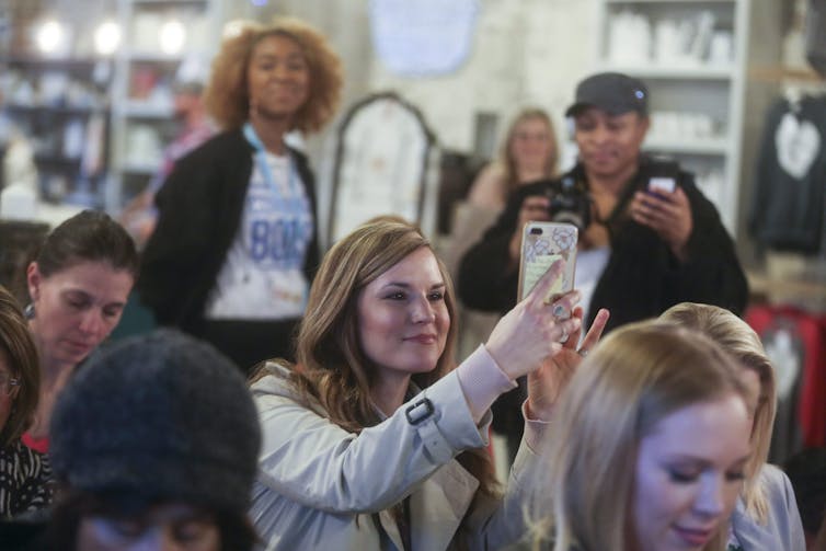 A woman with long hair in a crowd of other women takes a photo with her phone.