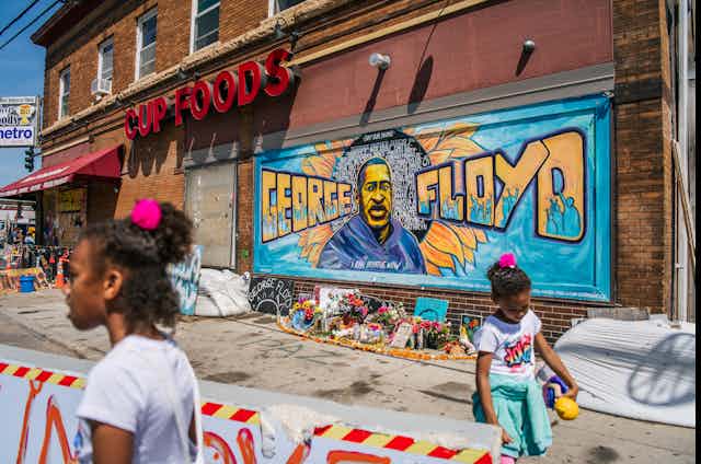 Two Black girls stand in front a mural for George Floyd.