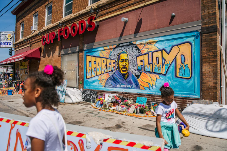 Two Black girls stand in front a mural for George Floyd.