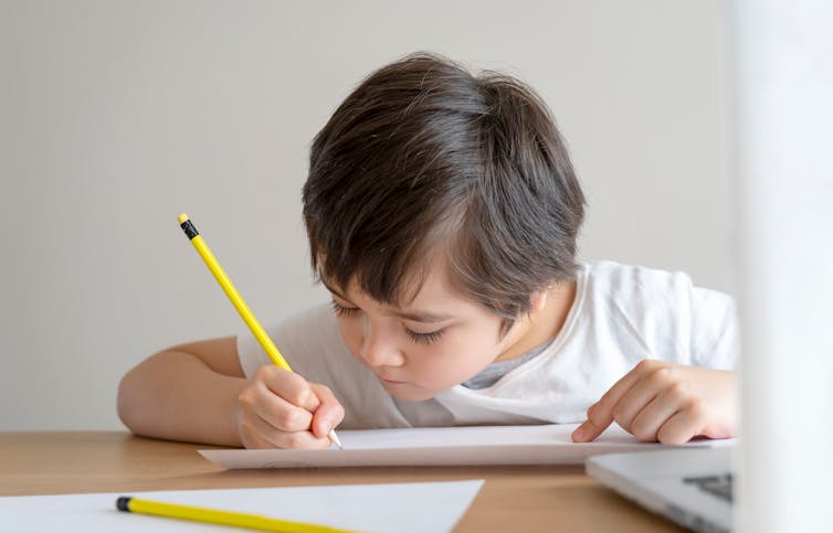 Primary school student writing on notepad with laptop open.