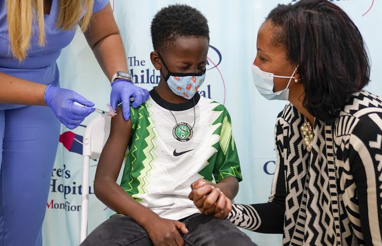 A Black woman in a face mask holds her son's hand as a health-care worker gives him an injection