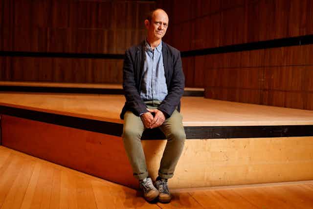 A balding man sits on a low wooden stage looking wryly at the camera.
