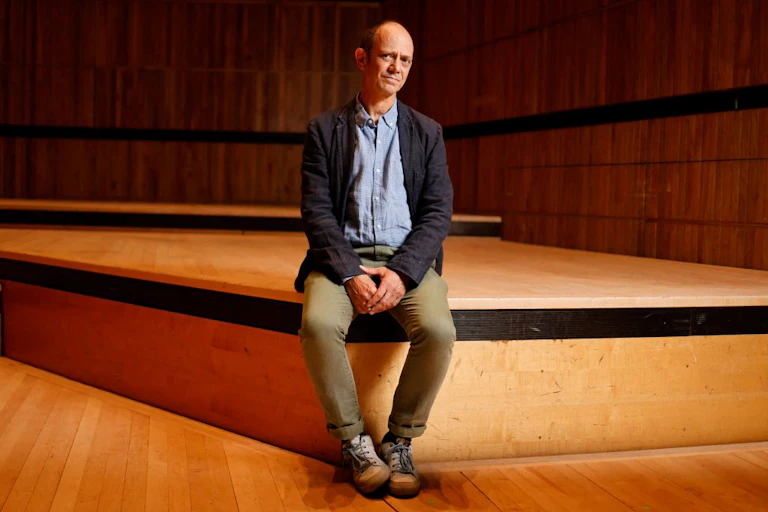 A balding man sits on a low wooden stage looking wryly at the camera.