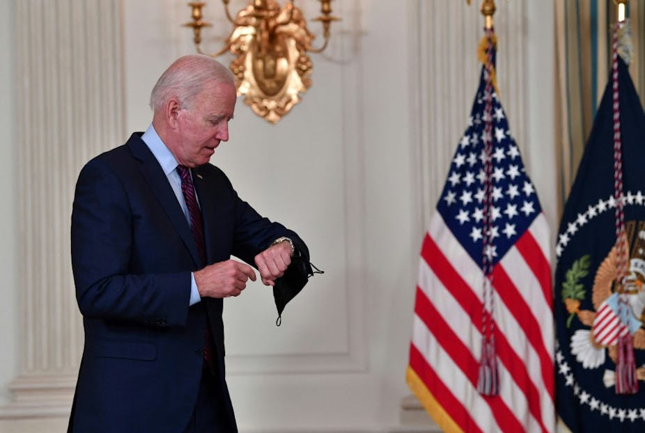Joe Biden regarde sa montre devant des drapeaux américains.