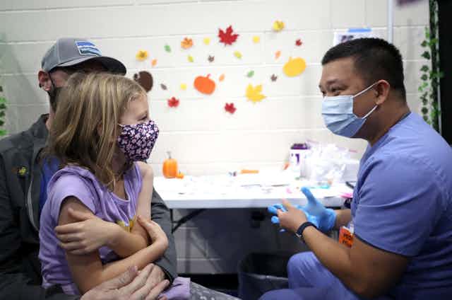 Discussion entre une petite fille, sur les genoux de son père et la personne qui va la vacciner.