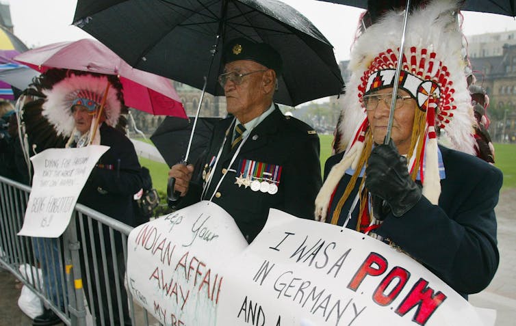 Indigenous veterans stand with placards protesting.