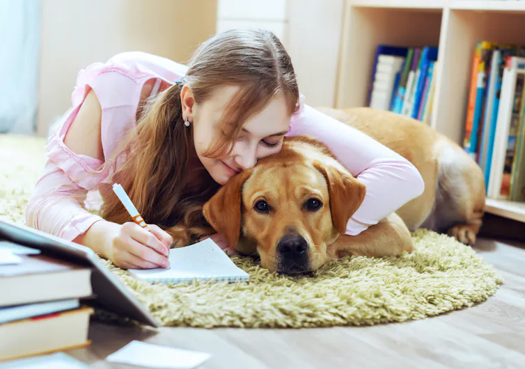 A student lying on the floor cuddles up with her dog as she takes notes