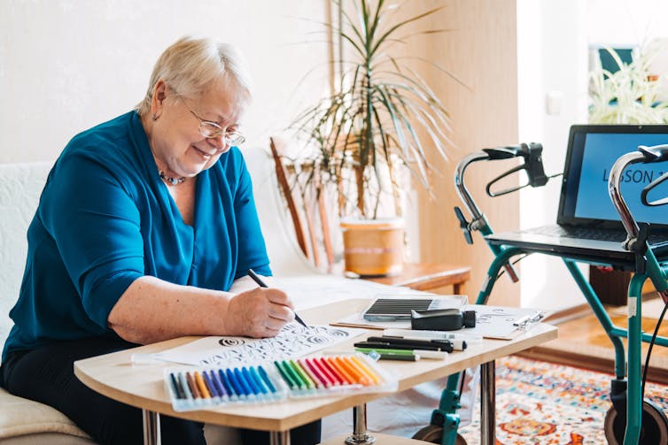 An older woman draws, a tablet next to her propped up on a walker