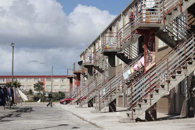 A man walks across a parking lot of a bleak housing project, heading to rows of metal stairs.