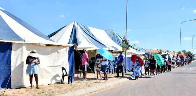 People line up along the road outside blue and white tents