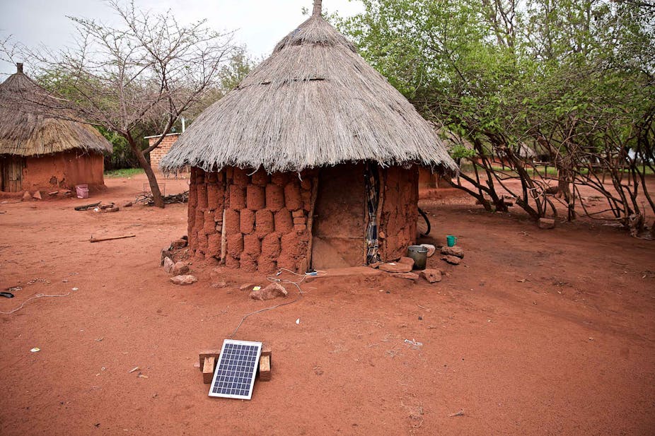 A single mud hut with a thatch roof attached by a wire to a single solar panel out in front of it in a village on Lake Kariba in Zimbabwe.