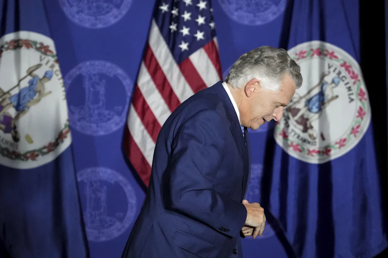 Virginia Govenor Terry McAuliffe looking dowcast walks off a stage with an American flag draped behind him.