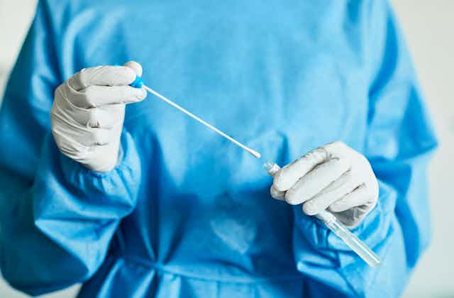 A medical professional in blue scrubs holding a swab with a small tube.