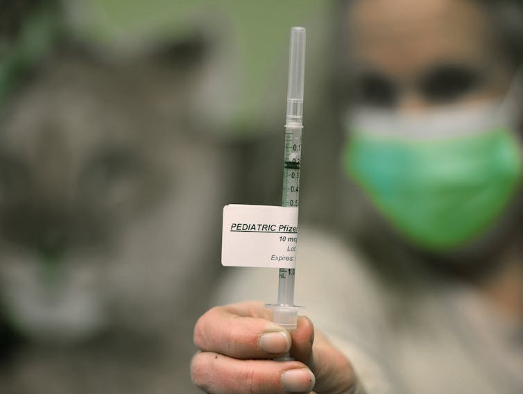 A syringe with a label reading 'PEDIATRIC Pfizer' in the foreground, held by a person in a mask, out of focus in the background