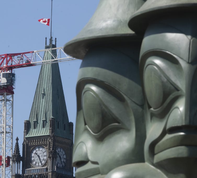A large bronze statue showing three united figures is seen in front of a background of Canadian flag at half mast and a construction crane.