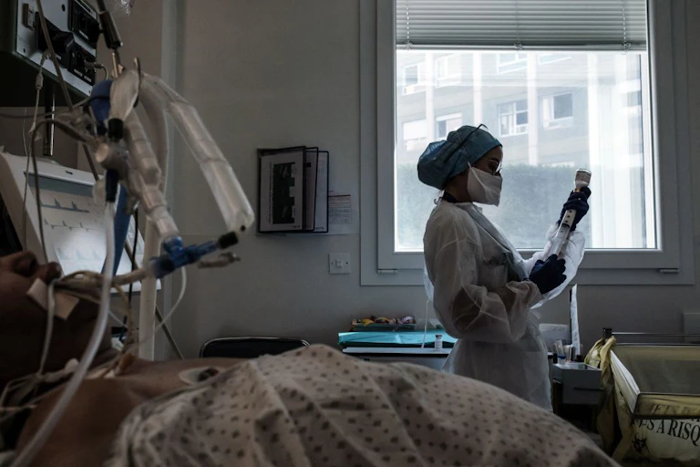 Nurse in PPE drawing liquid into syringe at a patient's bedside.