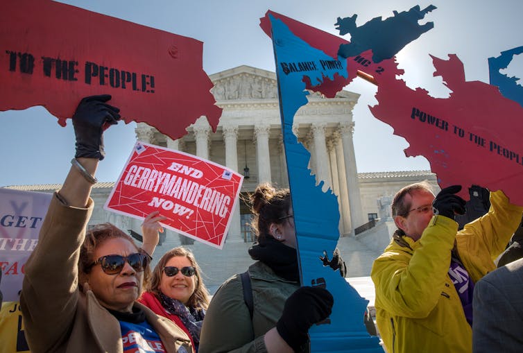 Four demonstrators hold signs opposed to gerrymandering with the Supreme Court building in the background.