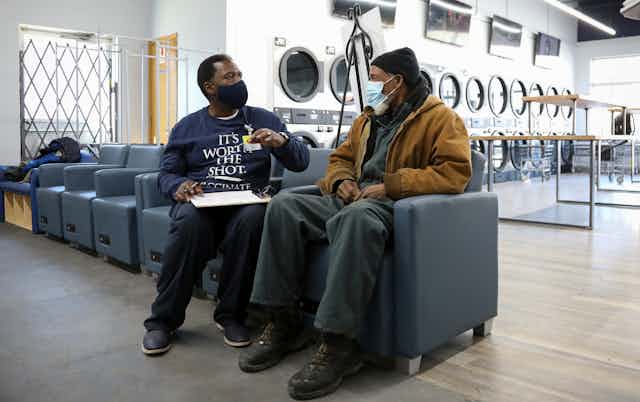Two seated men in a laundromat, both wearing face masks, one gesturing while holding a clipboard on his lap