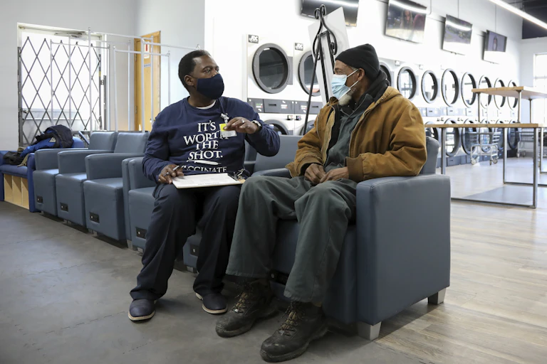 Two seated men in a laundromat, both wearing face masks, one gesturing while holding a clipboard on his lap