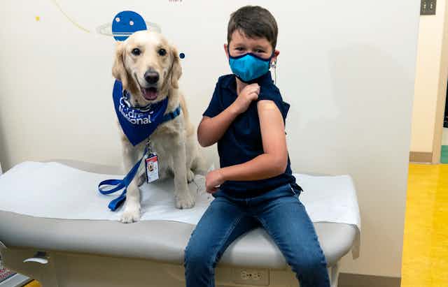 A boy sits on an examination table showing his vaccination site, with a golden retriever beside him."