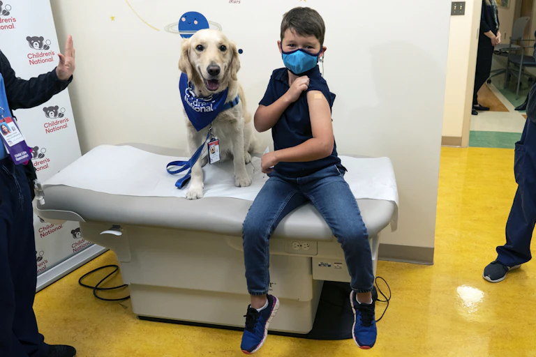 A boy sits on an examination table showing his vaccination site, with a golden retriever beside him."