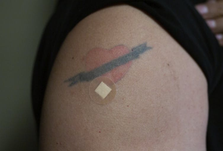 Un bras portant un tatouage de cœur et un petit pansement rond sur un point d’injection