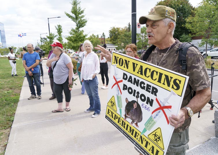 A man in camouflage T-shirt and hat holding an anti-vaccine sign in the foreground, with a group of people in the background