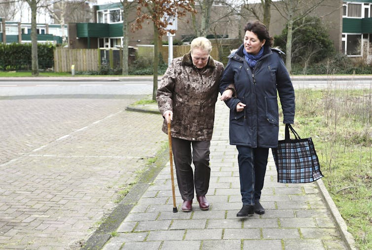 A caregiver helps an elderly lady walk along the pavement.