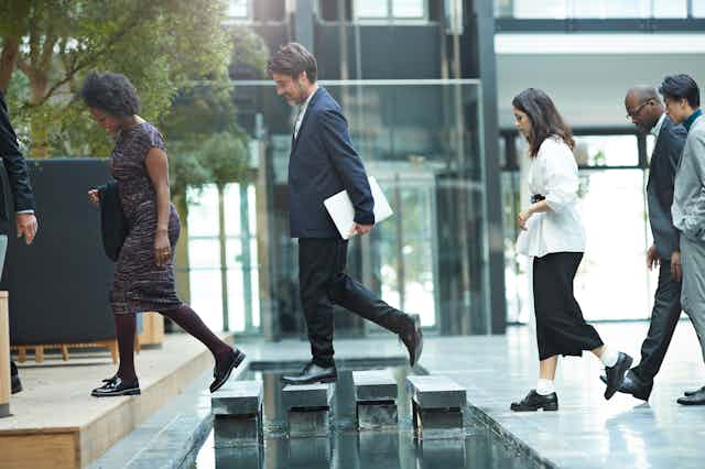 A line of employees in office attire walk over a stepping stones.