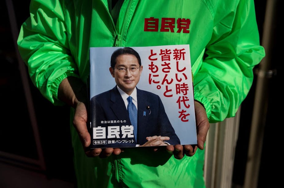 Un homme en imperméable vert tient un prospectus à l'effigie de  Kishida Fumio