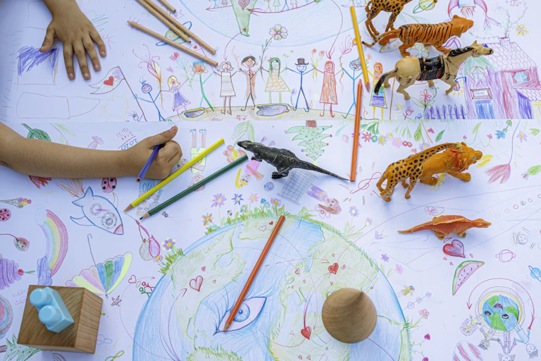 Childs hand holding colored pencil on top of large drawing of Earth, people holding hands and other illustrations