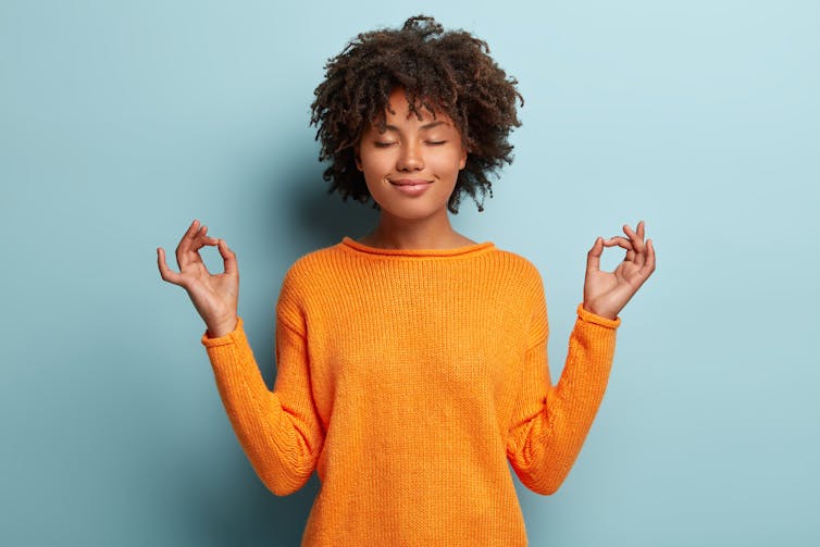 A Black woman wearing an orange sweater and her hair natural stands in front of a blue background with her eyes closed looking zen.