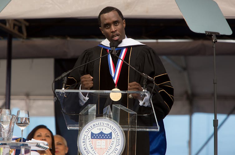 A Black man in a doctoral robe gives a speech on stage.