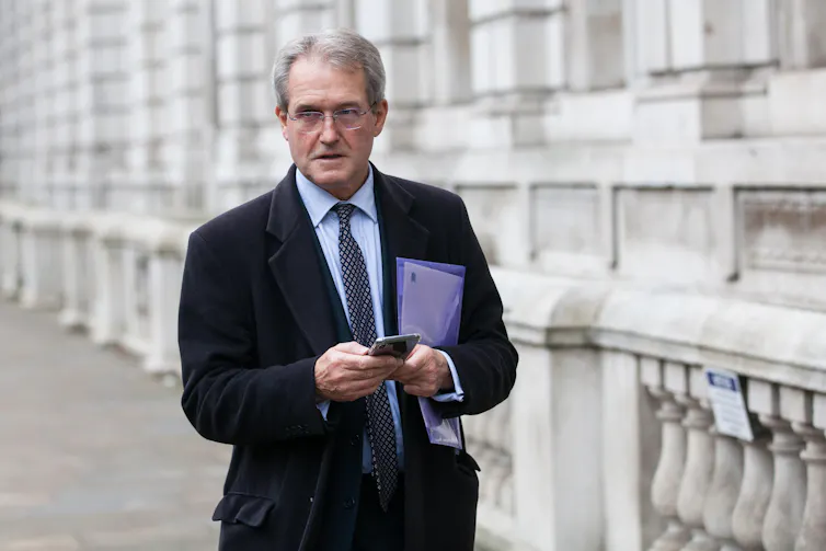 Owen Paterson walking in Whitehall, holding his phone.