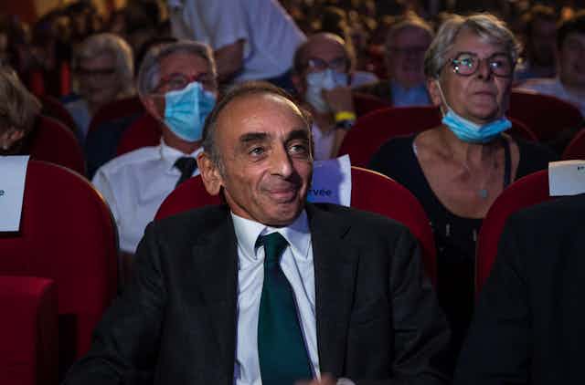 Eric Zemmour sitting in movie-theatre-style seating with people in covid masks sitting in the row behind him.