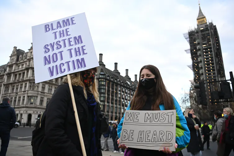 Two women protesting in front of the Westminster parliament. One is holding a sign reading 'blame the system, not the victim'.