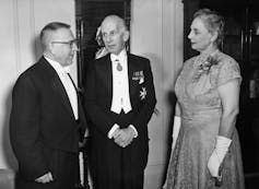 Two men in tuxedos chat as a woman in a dress and evening gloves looks on.