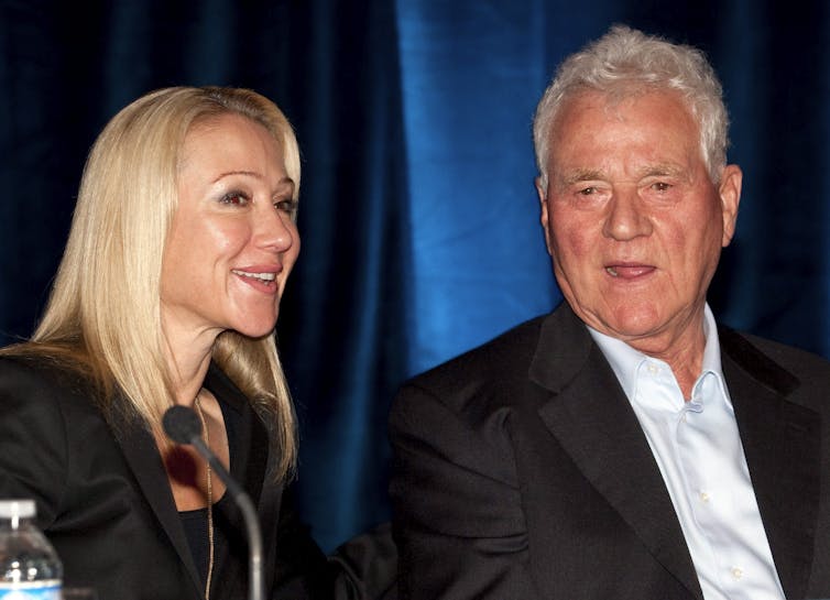 A blonde woman smiles while an elderly man talks to her.