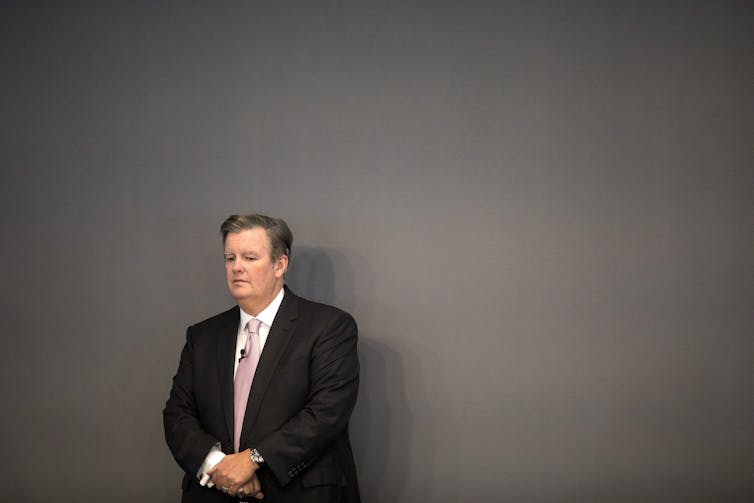 A man in a suit stands with his hands folded in front of him in front of a grey wall.