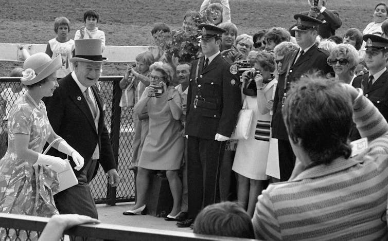 A man in top hat and tails escorts a woman in a flowered dress, hat, gloves and carrying a handbag past a crowd taking photos.